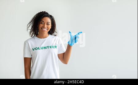Look here. Young african american volunteer in gloves pointing aside at empty space, standing over light studio wall Stock Photo
