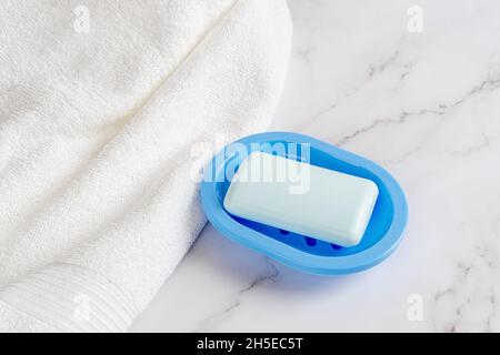 Light blue soap bar on a dish near white cotton terry towel over marble surface. Washing hands with soap, hygiene, purity and toiletries concepts. Stock Photo