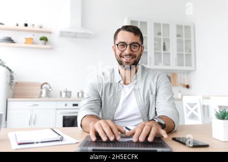 Smiling mature caucasian man in glasses working at computer on kitchen interior Stock Photo