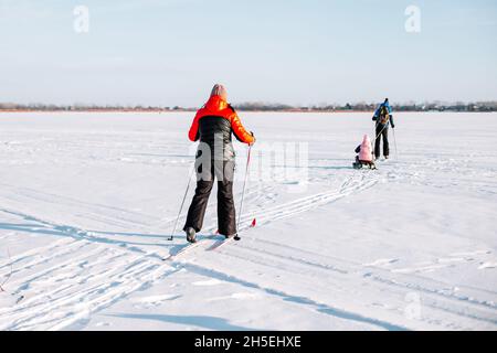 Family on ski trip. Young woman, man and child are skiing in winter on frozen river near forest, man is sledding child Stock Photo