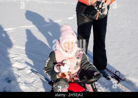 Little girl enjoys walk. Child squints from sun and enjoys sledge ride through snowy forest, shadows on snow Stock Photo