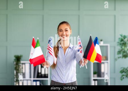 Learn foreign languages. Happy female teacher holding bunches of international flags, smiling to camera in office Stock Photo