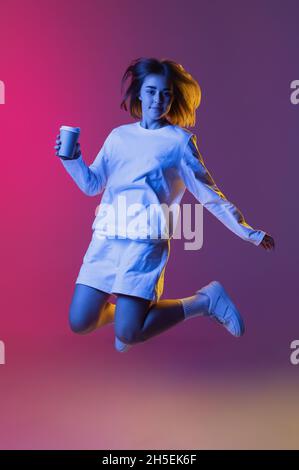 Studio shot of young cute girl, student in casual wear jumping isolated on pink-purple gradient studio backgroud in neon. Stock Photo