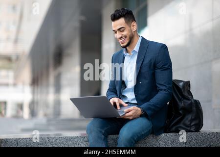 Positive middle eastern guy sitting on the street, using laptop Stock Photo