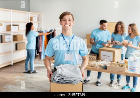 Portrait of male volunteer in uniform holding cardboard box with clothes for donation, smiling at camera Stock Photo