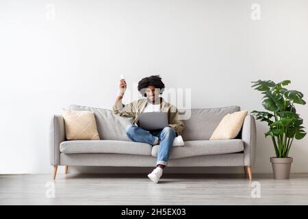 Young Black Man Sitting On Couch With Laptop And Using Remote Controller Stock Photo