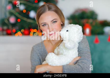 Portrait of a Beautiful Female Hugging Cute Little White Teddy Bear. Spending Christmas Holidays Near Christmas Tree at Home. Cozy Festive Evening. Stock Photo