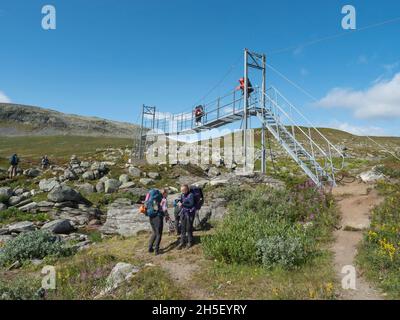 Sweden, Jokkmokk, Norrbotten, 4 August, 2021: Hikers crossing iron suspension foot bridge over river stream at Kungsleden hiking trail in Sweden Stock Photo