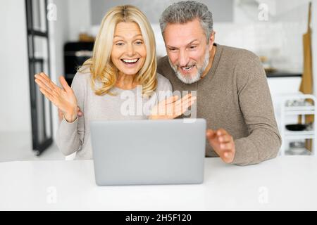 Shocked middle-aged couple staring at the laptop screen, surprised with unexpected news or email looks at the computer sitting at the table in kitchen at home. Mature spouses spends time online Stock Photo