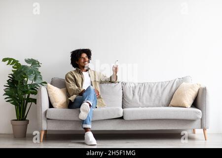 Happy Black Guy Sitting On Couch, Operating Air Conditioner With Remote Controller Stock Photo
