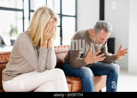 Home abuse concept. Depressed mid-aged woman sits on the couch and crying, an irritated and angry mature man is screaming on the background. A couple is arguing at home Stock Photo
