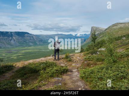 Man hiker admiring view on meandering glacial Rapadalen river delta valley at Sarek national park, Sweden Lapland with big rock, mountains and birch Stock Photo