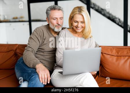 Middle-aged couple spends leisure time with a laptop at home. Spouses sit on the couch, watching video, web surfing or shopping online together, enjoying time online Stock Photo