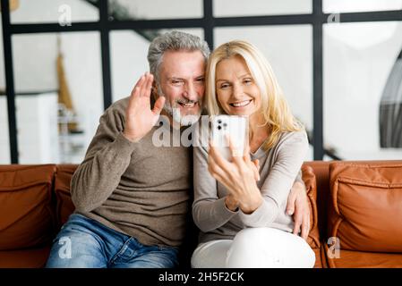 Middle-aged couple is taking selfie by a smartphone or making video call sitting at sofa in cozy living room. Happy spouses talking online with friends or family, using mobile app for video connection Stock Photo