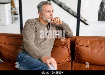 Portrait of thoughtful mid adult man sitting on the leather sofa at home. Handsome grey-haired man in casual wear relaxing on couch. Handsome mature bearded man looking away Stock Photo