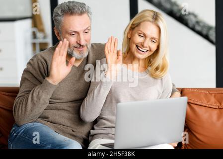 Middle-aged couple is using laptop for video call sitting on comfortable sofa in cozy living room. Cheerful spouses waving into webcam, involved virtual meeting with family. Online communication Stock Photo