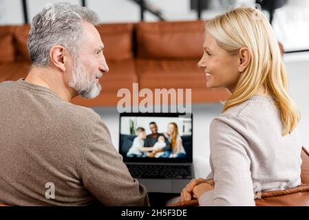Middle-aged couple using laptop computer for video connection with a grown daughter and her multiracial family, grey-haired man and woman talking online with family on computer screen Stock Photo