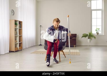 Man with broken leg and arm struggling with pain but trying to walk with crutch at home Stock Photo