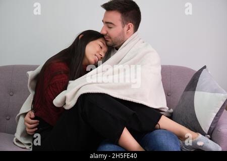 Young romantic couple covered with a blanket, hugging sitting on the couch. Stock Photo