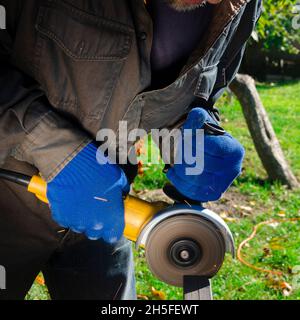 Close-up on the sides fly bright sparks from the angle grinder machine. A young male welder in a white working gloves grinds a metal product with angl Stock Photo