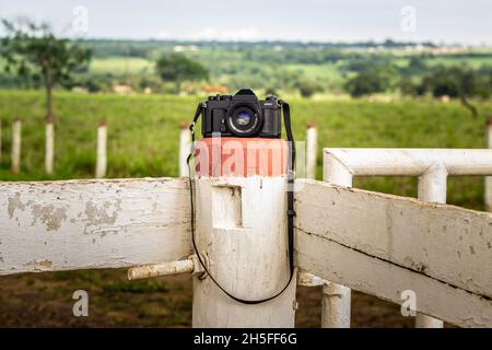A vintage film camera over a white-painted fence with brown accents and countryside in the background.