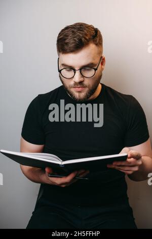 Portrait of caucasian bearded man wearing glasses on white background, reading book. Holding an open copybook, looking down. White wall. Stock Photo