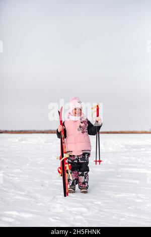 Child is enjoying winter walk. Happy little girl is standing on road covered with snow, holding children skis in her hands and rejoicing at her Stock Photo