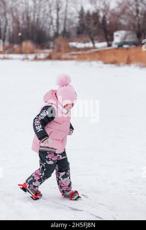 Little girl on walk. Happy little girl in pink warm suit walks on snowshoes on snowy road near bank of frozen river, side view Stock Photo