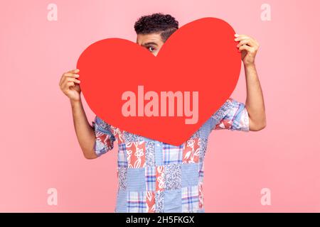 Positive man with dark hair hiding face behind big red heart and looking at camera with curious prying eyes, feeling affection fondness. Indoor studio shot isolated on pink background. Stock Photo