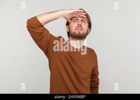 Portrait of serious concentrated attentive man with beard wearing sweatshirt, standing with hand on forehead and looking for something. Indoor studio shot isolated on gray background. Stock Photo