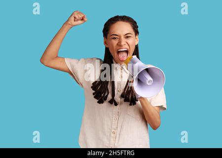 Portrait of woman with black dreadlocks standing with raised hands and holding megaphone, screaming in loud speaker, protesting, wearing white shirt. Indoor studio shot isolated on blue background. Stock Photo
