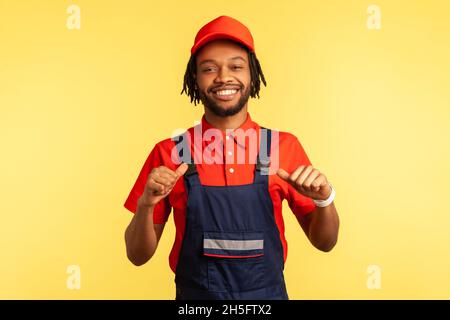 Smiling happy bearded courier or builder wearing overalls, looking at camera with proud expression, pointing at himself, satisfied of work done. Indoor studio shot isolated on yellow background. Stock Photo