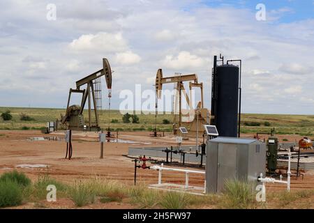 Two oil - gas well pump jack at drilling site with equipment and holding tanks on praries with low hill behind and blue cloudy sky Stock Photo