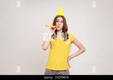 Portrait of happy adorable teenage girl in yellow T-shirt with birthday hat blowing party horn, having fun at holiday celebration, looks at camera. Indoor studio shot isolated on gray background. Stock Photo