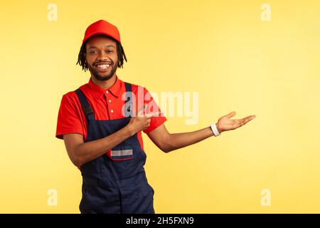 Smiling bearded mechanic wearing overalls presenting copy space on palm, pointing aside, advertisement of delivery or housekeeping service. Indoor studio shot isolated on yellow background. Stock Photo
