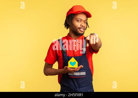 Good looking mover or handyman in red cap and blue overalls, holding paper house in hands and pointing finger to camera, offering home repair services. Indoor studio shot isolated on yellow background Stock Photo