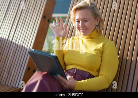 Smiling middle-aged caucasian woman having video call online on tablet, e-learning, tutoring, teaching remotely on distance in park. Stock Photo