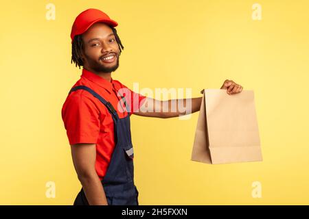 Side view portrait of happy professional courier wearing overalls giving groceries bag, delivering takeaway food, looking smiling to camera. Indoor studio shot isolated on yellow background. Stock Photo