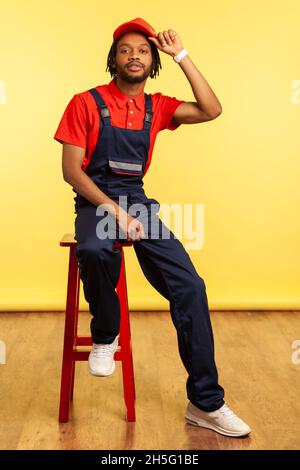 Portrait of handsome stylish male worker sitting on chair looking directly at camera, keeping hand on his visor cap. Indoor studio shot isolated on yellow background. Stock Photo
