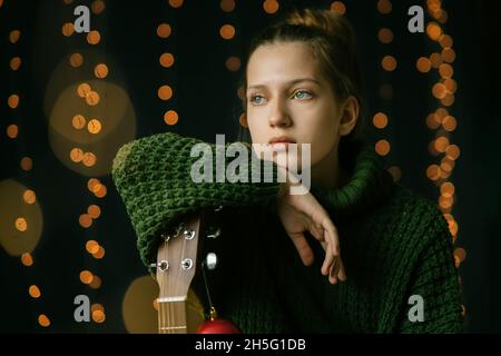 A beautiful teenager girl dreamily looks to the side, leaning on the neck of a guitar. Calm, serene face of a teenage girl, expressive eyes, a look to Stock Photo