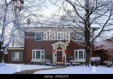 Beautiful brick house with bay windows with Christmas tree showing through and decorated pillars and sled on porch in snow framed by winter trees Stock Photo