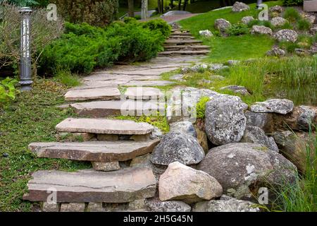 stone staircase with steps made of wild natural stone on slope of hill park landscape with bushes and trees, ground garden lantern among green plants Stock Photo