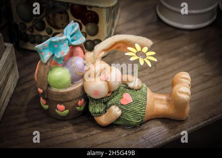 Ceramic Easter Decoratioin bunny asleep against a basket of eggs holding a flower - on shelf with other items Stock Photo