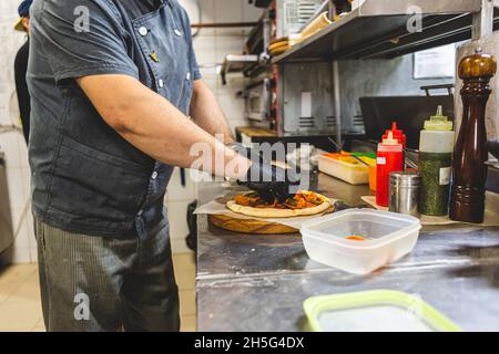 Closeup hand of chef baker in white uniform cutting pizza at kitchen Stock Photo