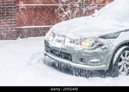 Closeup of front of silver car in snowstorm with lights on but covered in snow and icicles Stock Photo