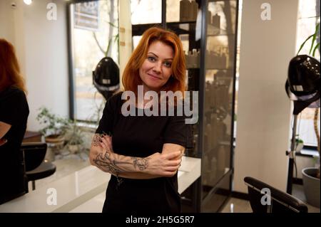 Confident portrait of a attractive fashionable redhead pretty woman hair stylist with tattooed arms, smiles looking at camera posing with crossed arms Stock Photo