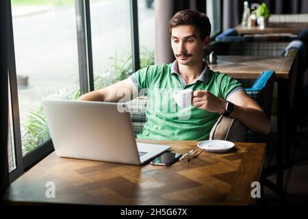 Portrait of relaxed calm handsome businessman wearing green T-shirt, enjoying hot coffee while working online on laptop, having break during work. Indoor shot near big window, cafe background. Stock Photo