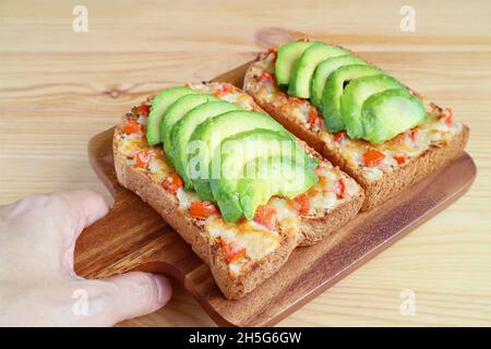 Hand holding a breadboard of grilled cheese toasts topped with tomato and sliced avocado Stock Photo