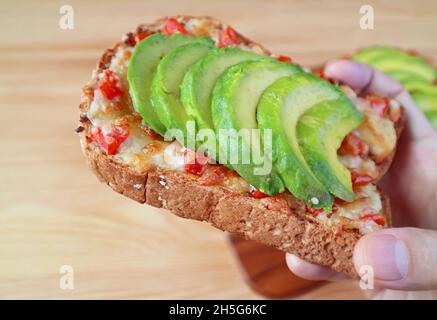 Closeup a delectable grilled cheese toast topped with tomato and sliced avocado in hand Stock Photo