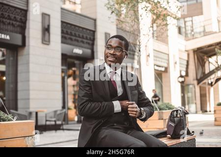 Portrait of black african american businessman in suit with glasses and headphones sits on the background of city buildings outdoors Stock Photo